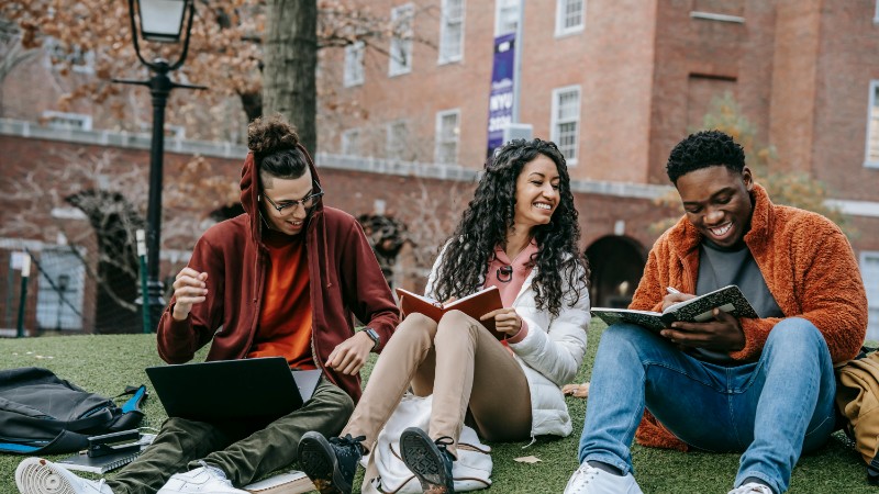 Full body of happy diverse students with notebooks and laptop sitting on grassy lawn on campus of university while studying together, tags: und - pexel